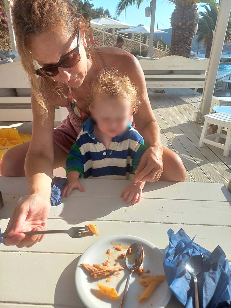 Babysitter helping a toddler eat during a meal at an outdoor café in Sifnos